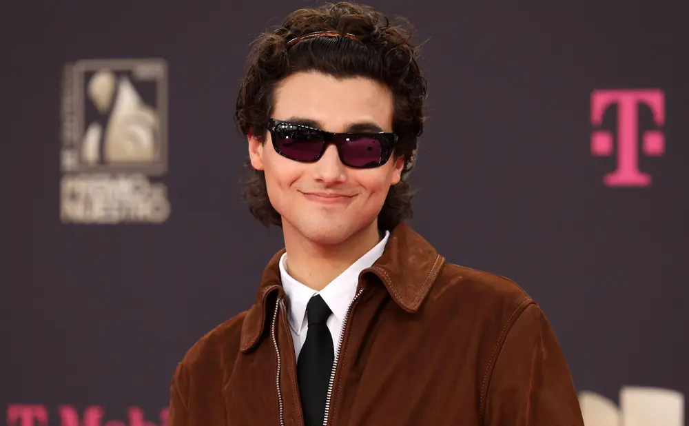 A young man with curly hair and sunglasses smiles at the camera, wearing a brown jacket and a black tie.