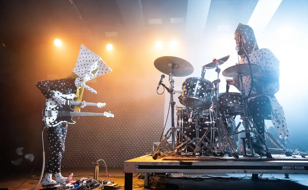 Two musicians in black and white polka dot outfits perform on stage. One plays a double-neck guitar, the other a drum kit.