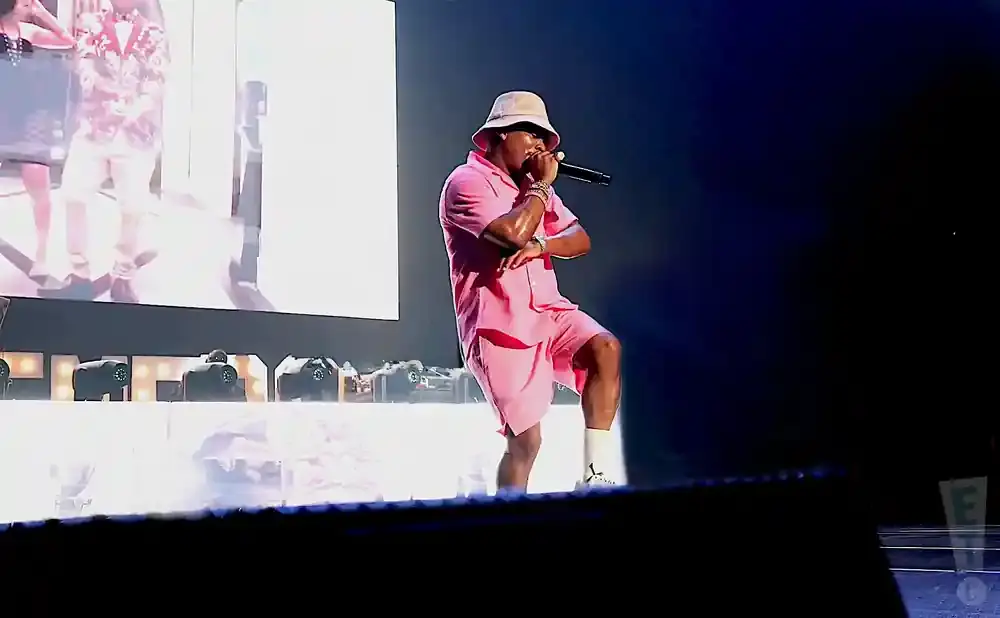 A rapper in a pink bucket hat and matching pink shorts suit performs on stage, holding a microphone.
