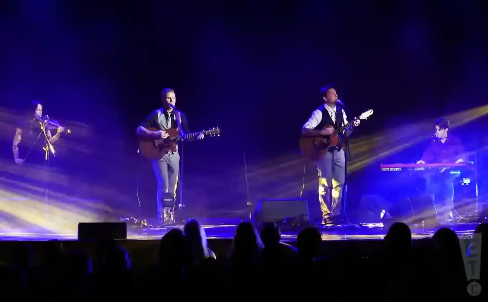 A band performs on stage with blue lighting, featuring a violinist, two guitarists, and a keyboardist.