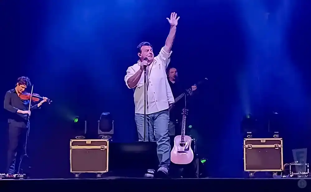 A male singer on stage with his arm raised, holding a microphone. A violinist stands to his left, and a guitarist is behind him.