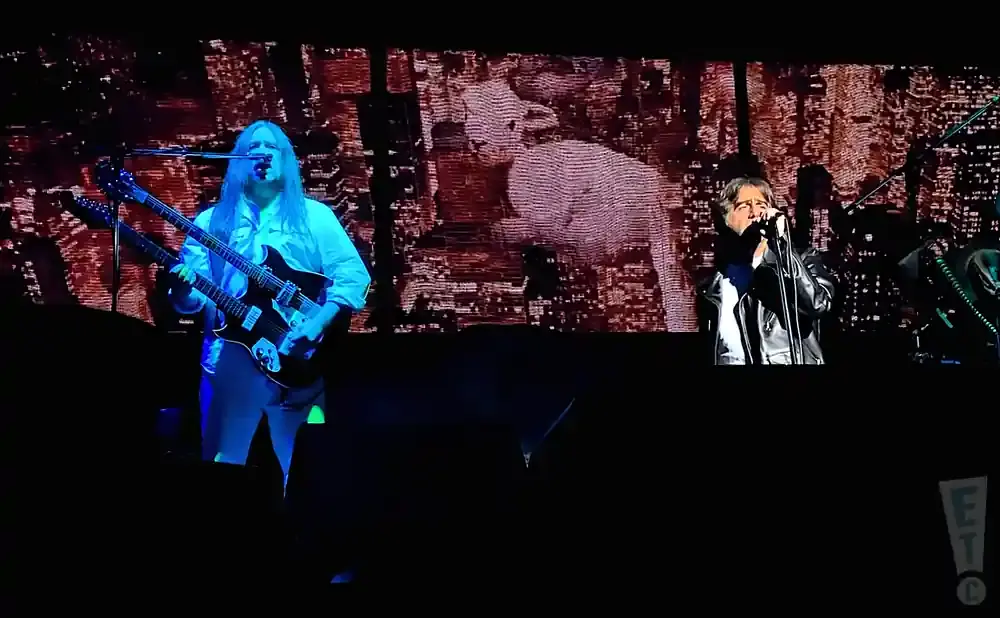 A man with long blond hair plays a double-neck guitar on stage.
