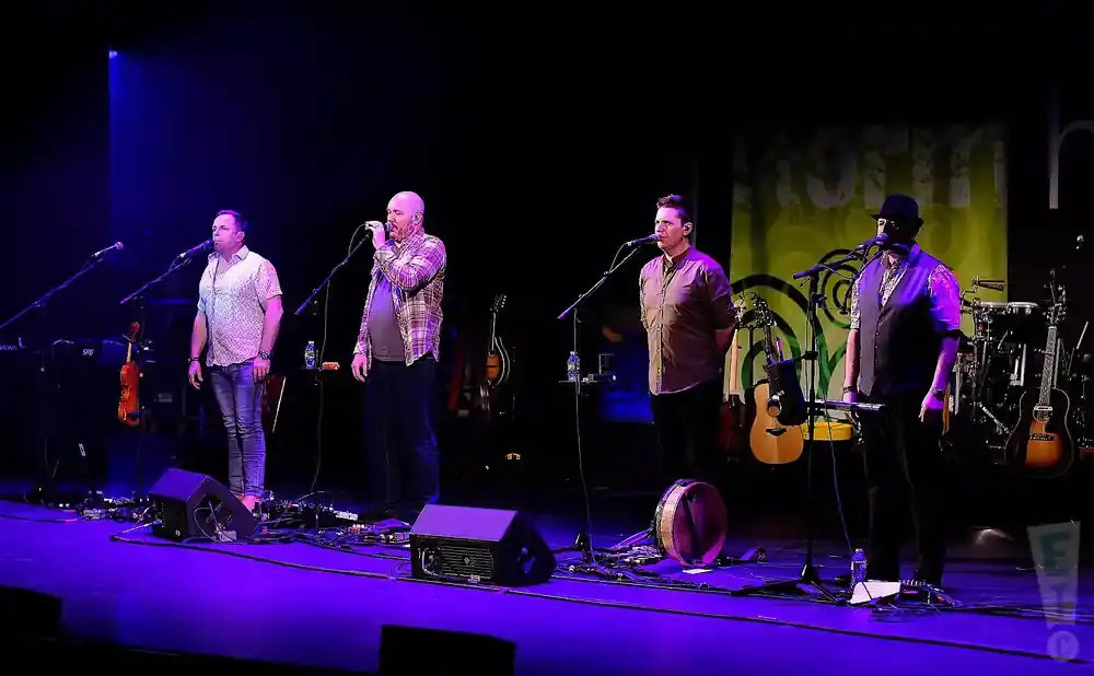Four men sing on a stage with instruments, including guitars and a drum.