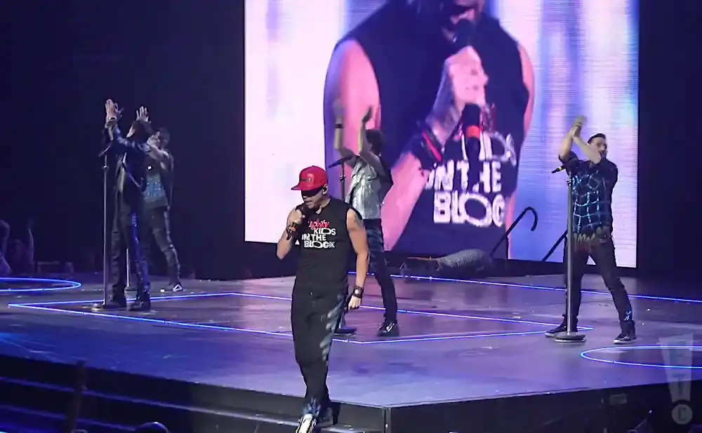 A group of five male singers perform on stage under bright lights, with a large screen behind them displaying a band member.