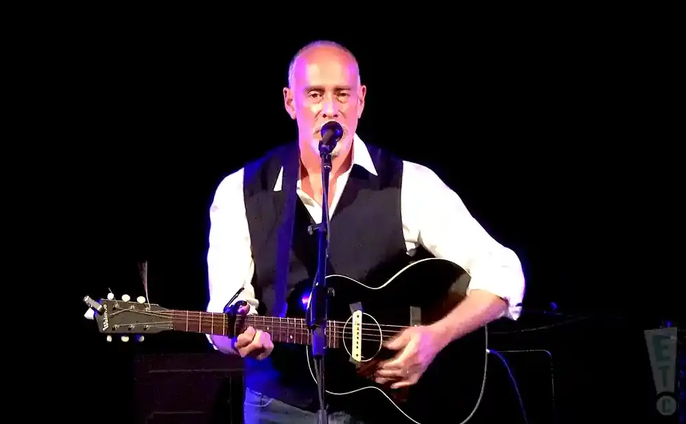 A bald man in a white shirt and vest plays an acoustic guitar and sings into a microphone.