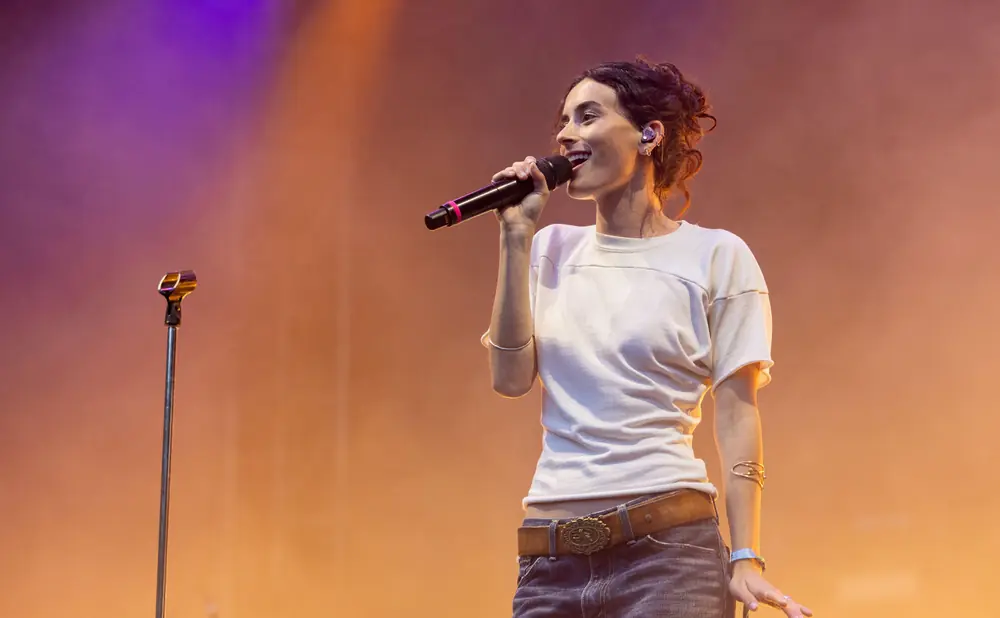 Singer on stage holds a microphone, wearing a white shirt, jeans, and a brown belt with an ornate buckle.