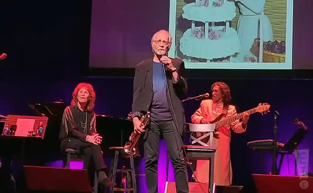 A man holds a saxophone while standing center stage; a woman sits left playing piano, and a man plays bass guitar right.