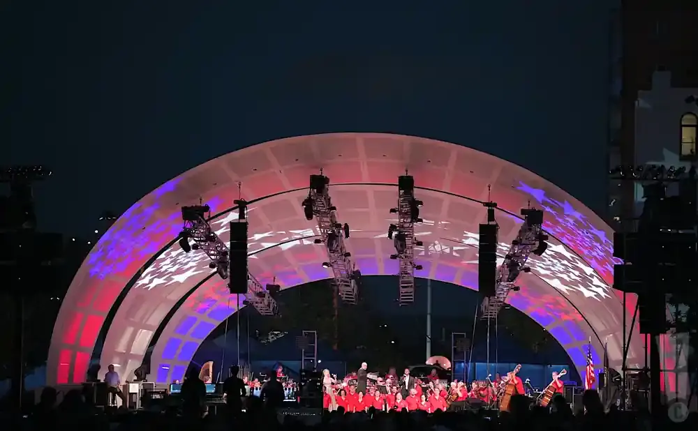 Outdoor orchestral performance on a stage with a large arched roof displaying red, white, and blue star patterns.