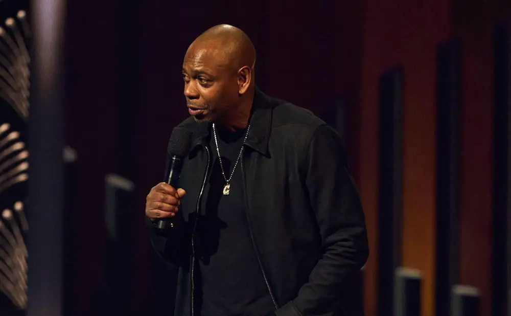 Dave Chappelle on stage holding a microphone and wearing a black jacket and necklace.
