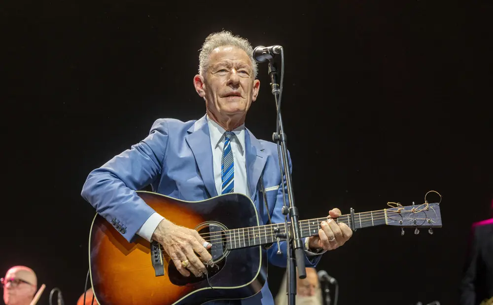 A man in a blue suit plays an acoustic guitar on stage, with a microphone in front of him.