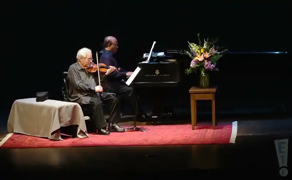 Two men play music on stage: one on violin, the other on a Steinway piano. A floral arrangement sits beside the piano.