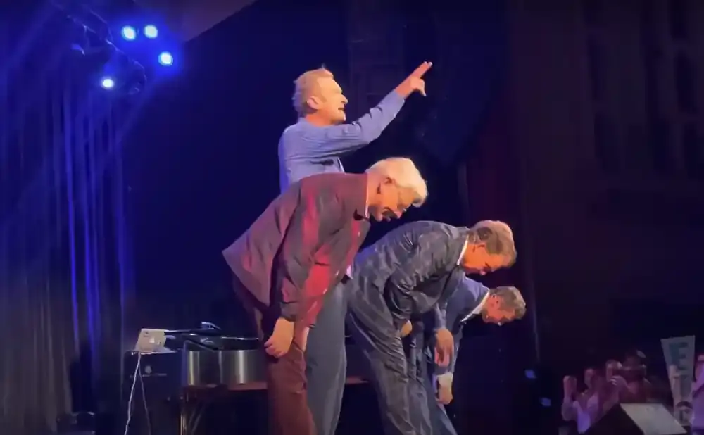Four men in suits bow on stage, with one man in a blue shirt standing behind them pointing.