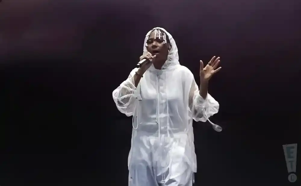 A performer in a white hooded outfit sings into a microphone, with a black background.