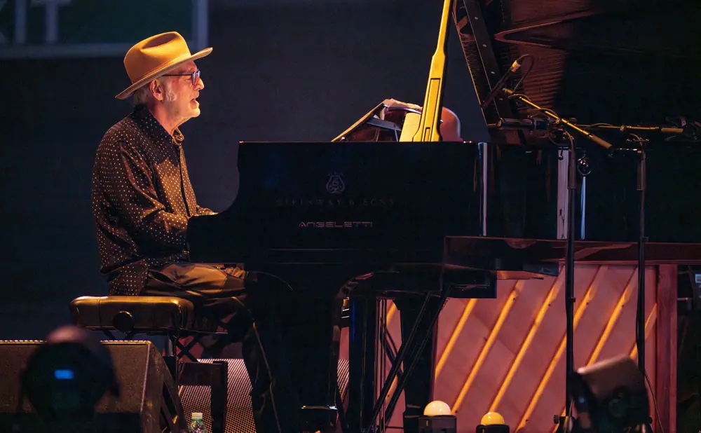 A man in a tan hat plays a Steinway piano on a dimly lit stage, with warm lights in the background.