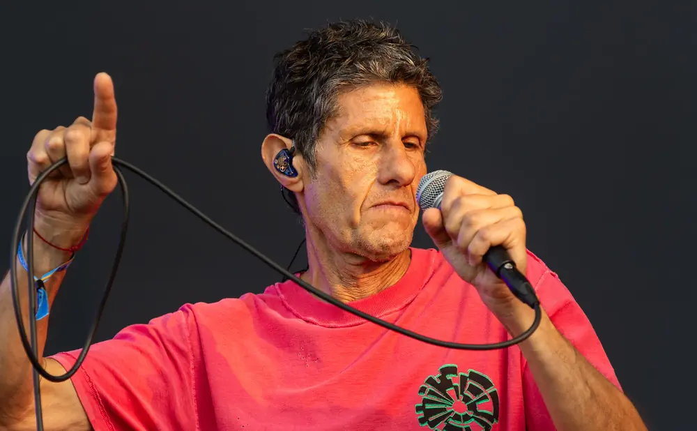 A man with grey hair sings into a microphone, wearing a pink shirt and an ear monitor.