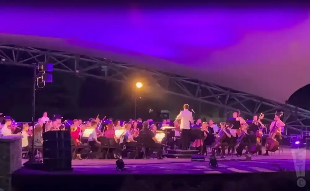 An orchestra performs under purple lights on an outdoor stage, with a conductor in a white suit.