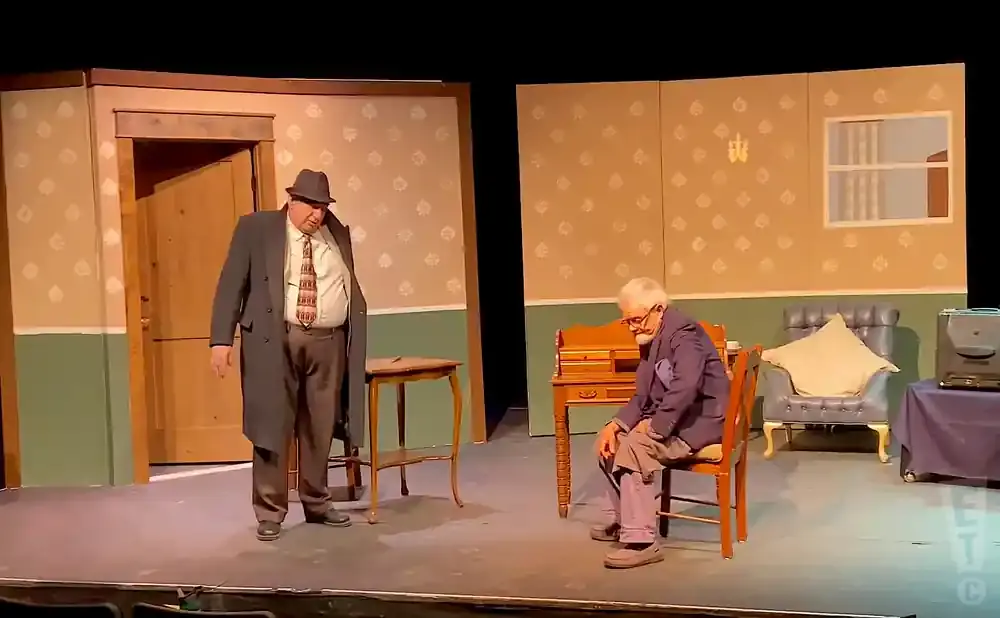 Two men are on a stage in a set. One man in a suit and hat stands near a door, the other man in a suit sits in a chair.