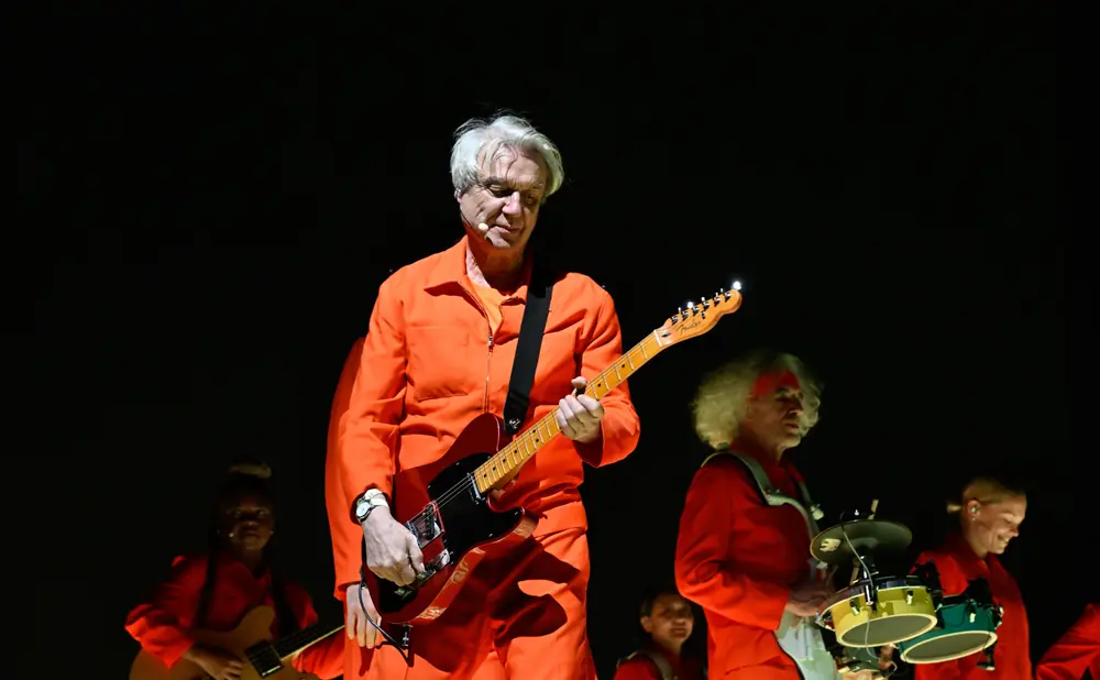 Musicians in orange jumpsuits perform on stage with David Byrne in the foreground playing a red guitar.