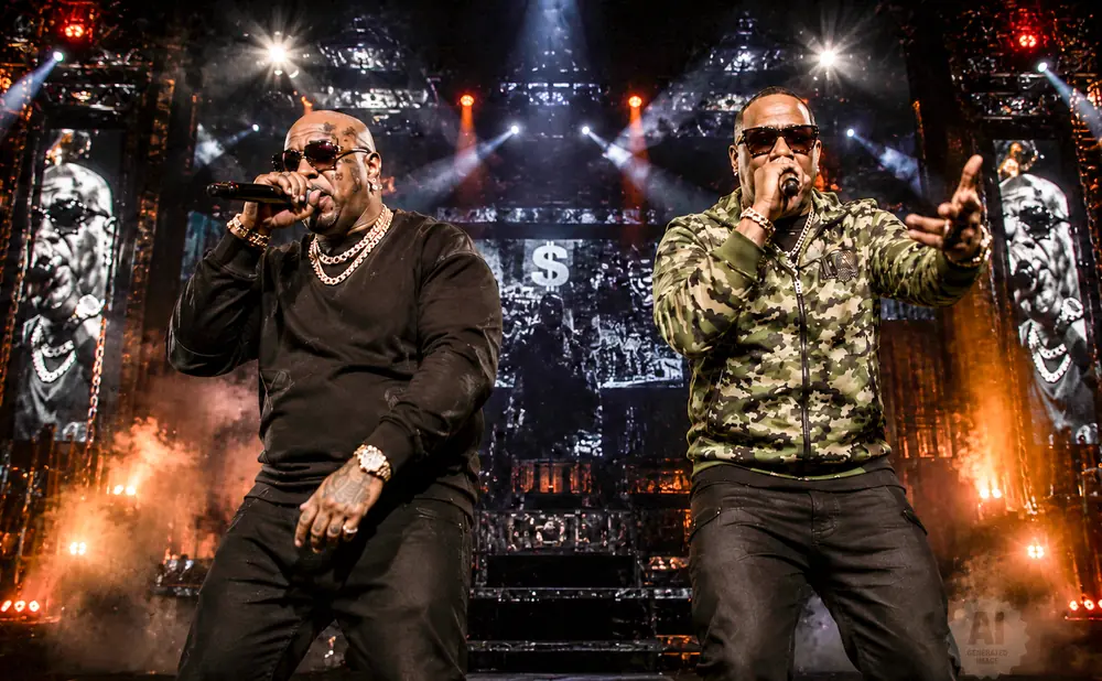 Two rappers in chains and sunglasses perform on stage with a large screen behind them showing their faces and dollar signs.