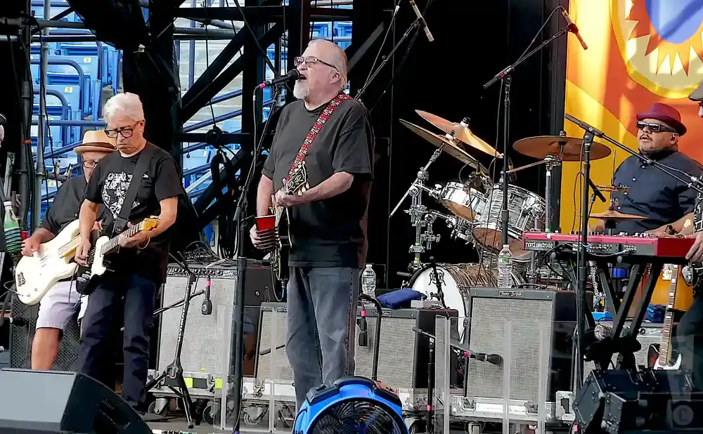 A band performs on stage. A guitarist in glasses wears a black t-shirt and jeans, playing a white electric guitar.