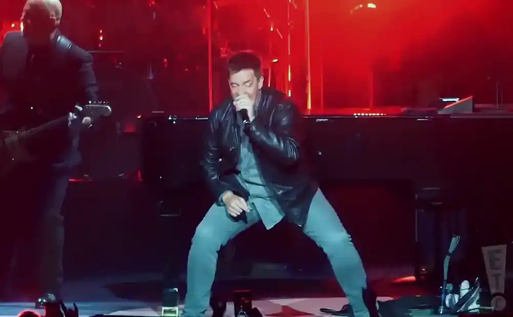 A male singer in a leather jacket and grey pants sings into a microphone on stage.