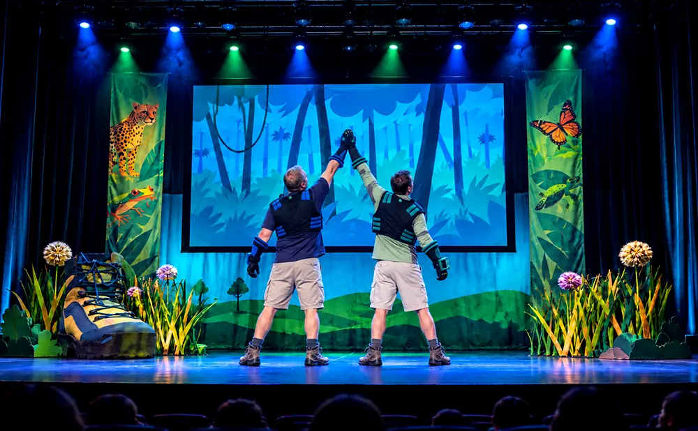 Two performers in safari outfits high-five on a stage with jungle-themed backdrops and props.