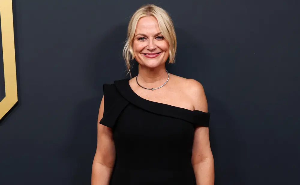 A blonde woman in a black off-the-shoulder dress smiles warmly at the camera.