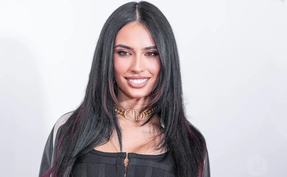 A woman with long dark hair with pink streaks smiles, wearing a black ribbed top and a gold 'CD' choker.