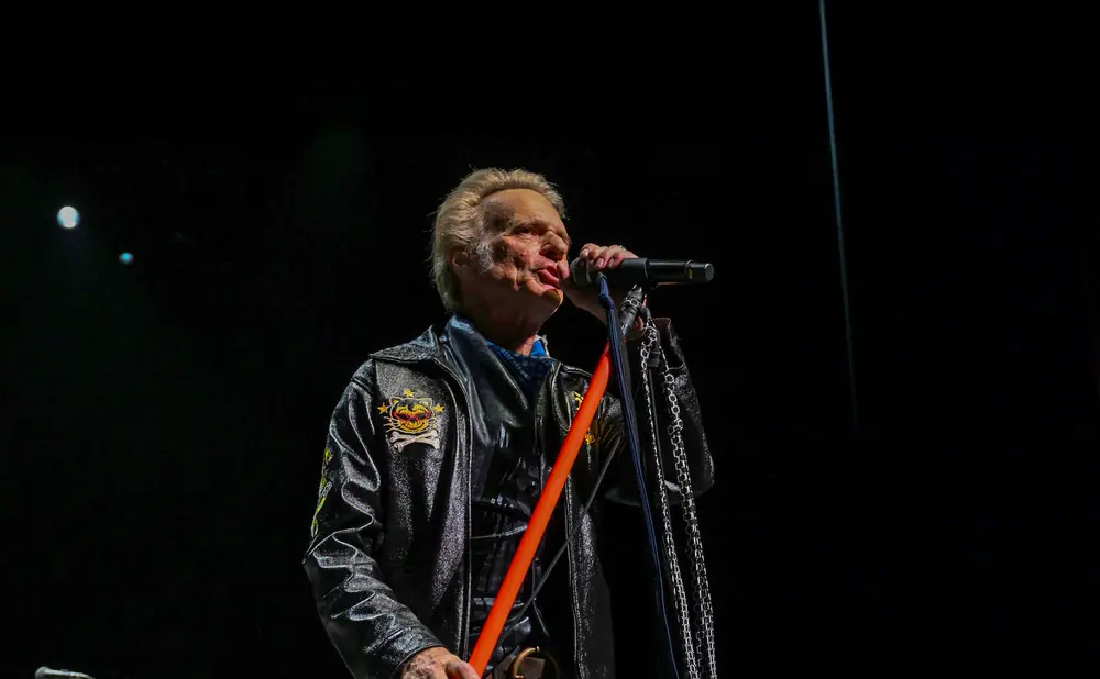 A man in a black leather jacket sings into a microphone with chains hanging from his jacket.