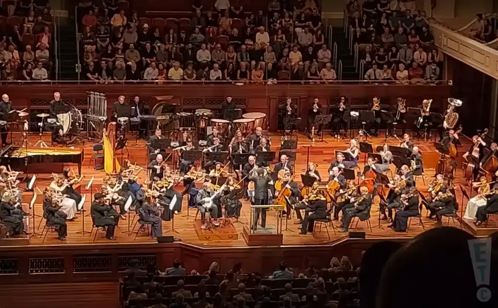 An orchestra performs on stage with a conductor in front, and an audience in the background.