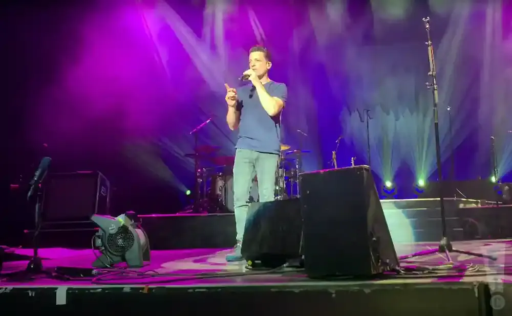A man sings into a microphone on a stage with purple and green lights.