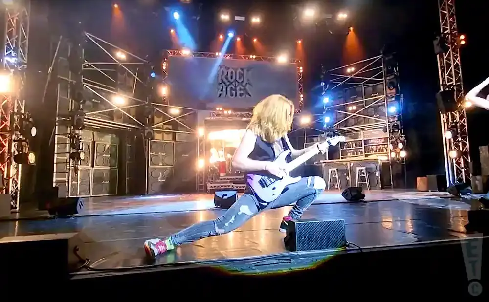 A guitarist on stage in a low lunge position, playing a white electric guitar. The backdrop features stacked amplifiers and a 