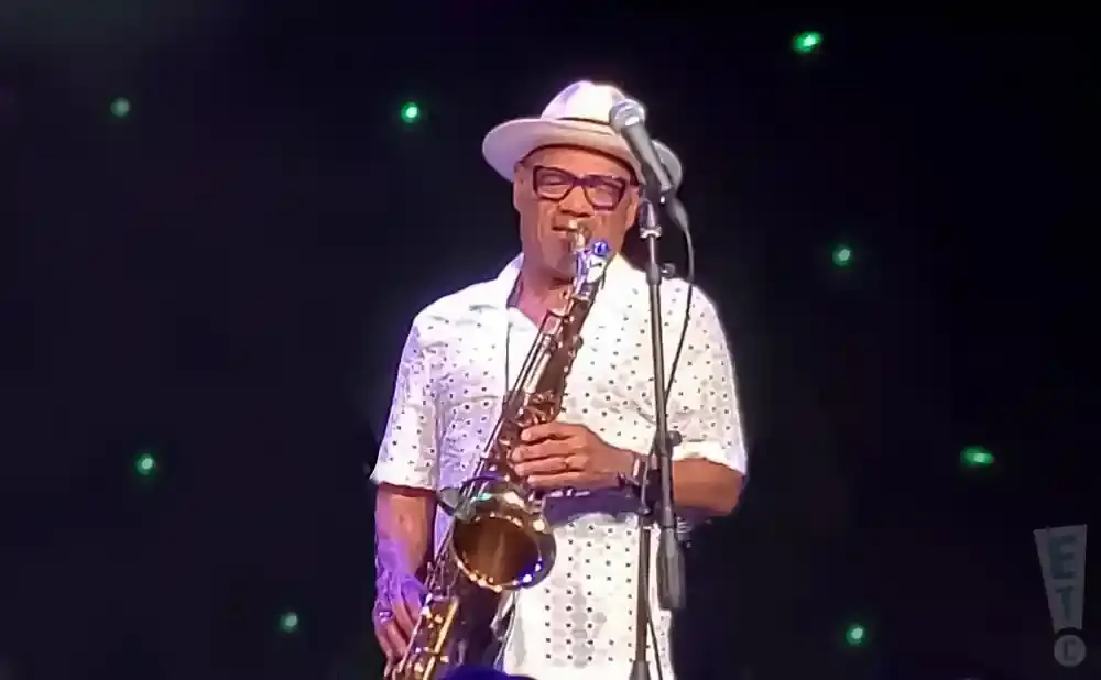 A Black man in a white hat and patterned shirt plays a saxophone on stage with green lights in the background.