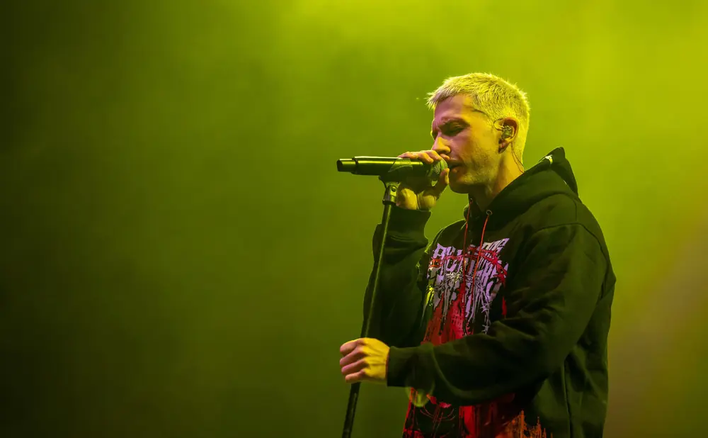 A singer with bleached hair performs on stage, wearing a black hoodie with a red graphic.