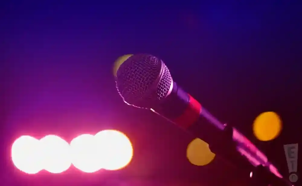 A microphone on a stand is lit by purple and pink stage lights, with blurred yellow and white circles in the background.