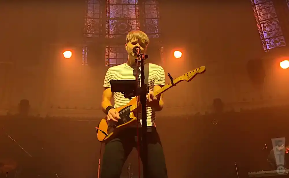 Man in a striped shirt plays a yellow electric guitar on stage with warm lighting.