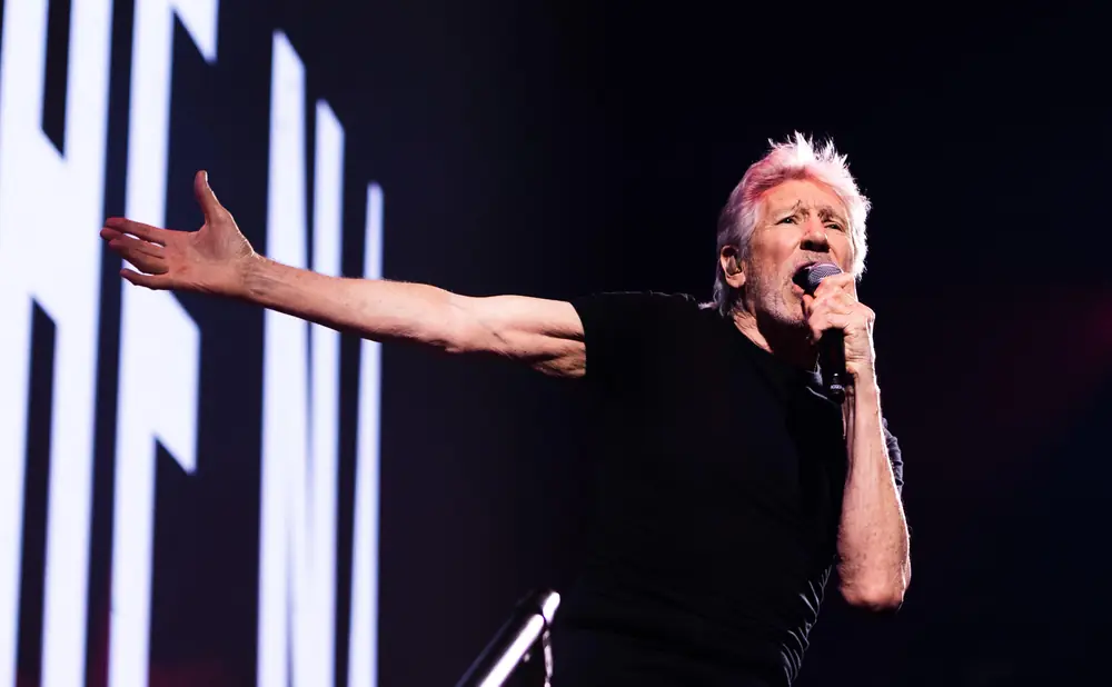 Roger Waters performs on stage with his arm outstretched and holding a microphone.