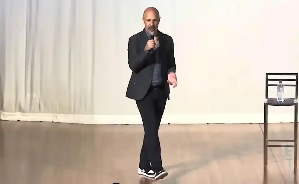 A bald man in a suit speaks into a microphone, while standing on a wooden stage.