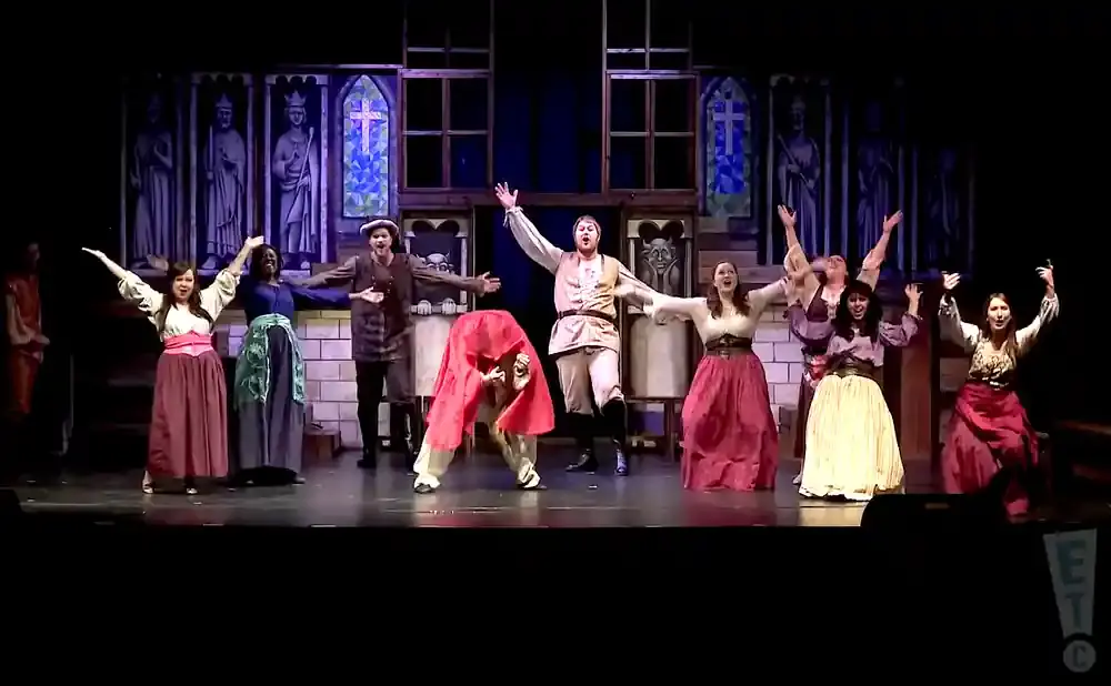 A group of actors in costume perform on a stage with a medieval backdrop.