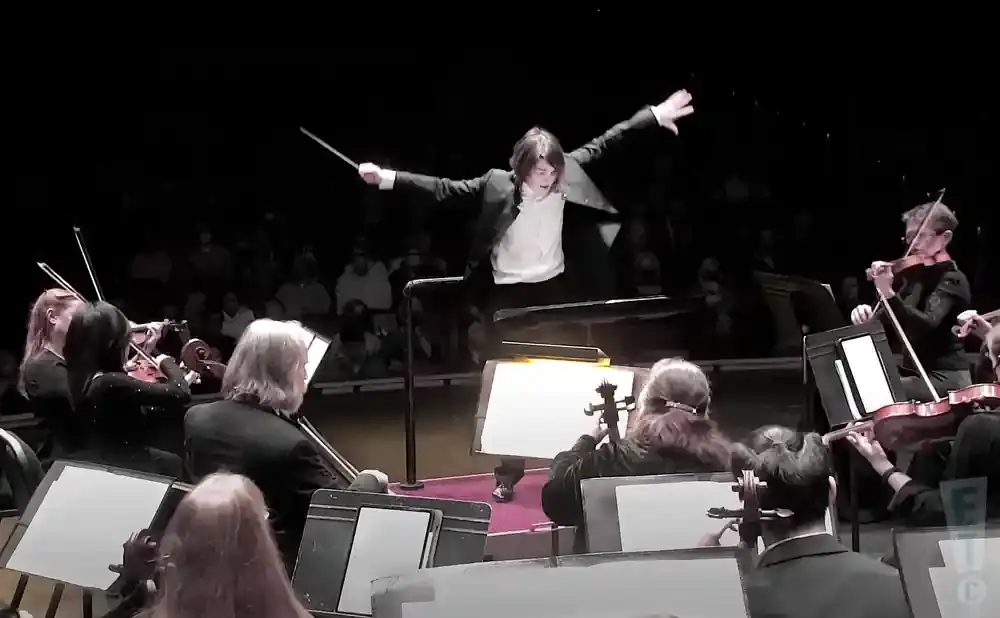 An orchestra conductor directs musicians in a dark theater.