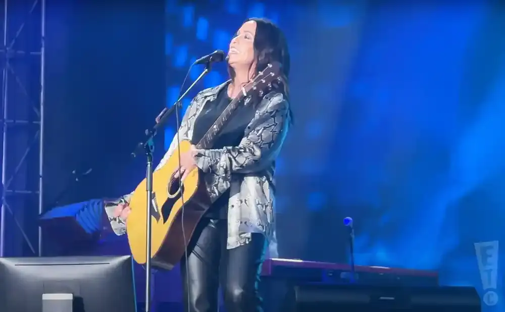 A woman with dark hair plays an acoustic guitar on a stage with blue lighting.