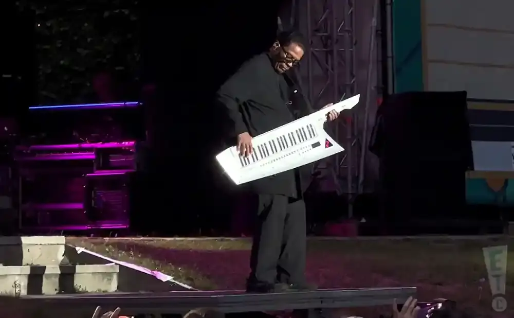 A man in a black outfit plays a white keytar on a stage with purple lighting.