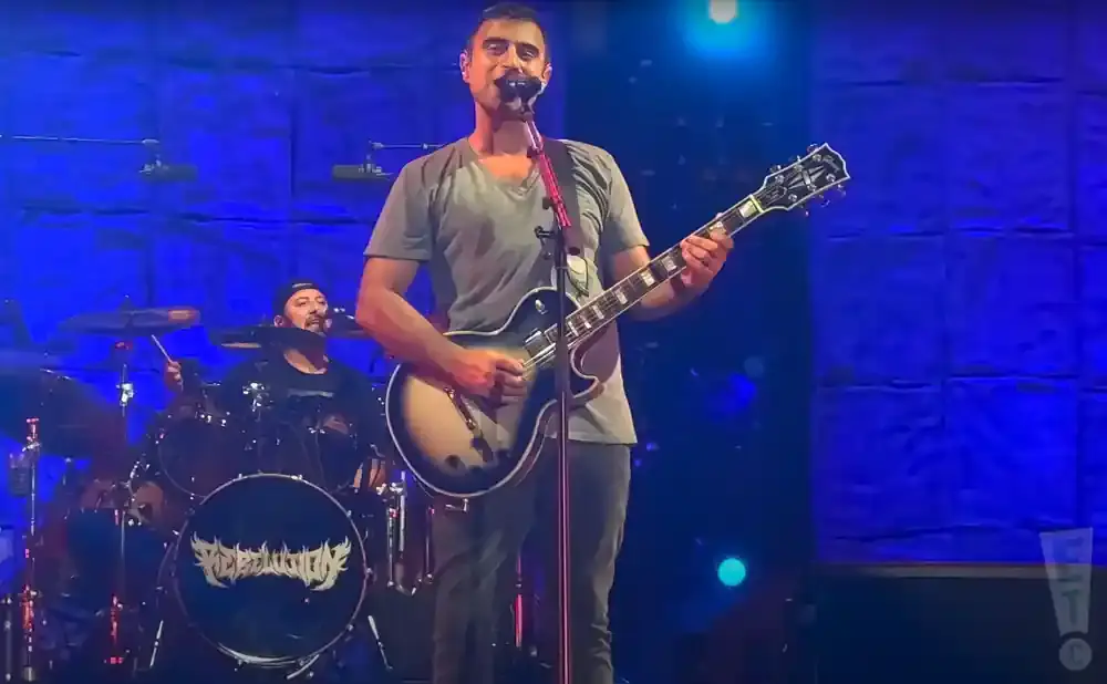 A male guitarist sings and plays a guitar on stage with a drummer in the background.