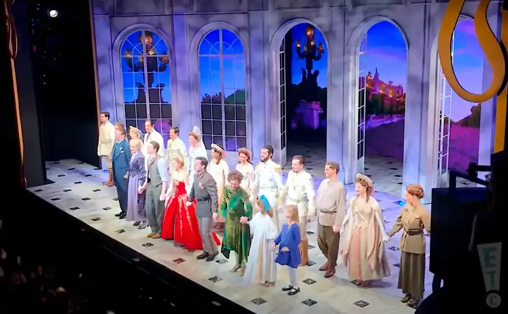 A cast of actors in historical costumes stand on a stage in front of an elaborate set depicting a garden with arched windows.
