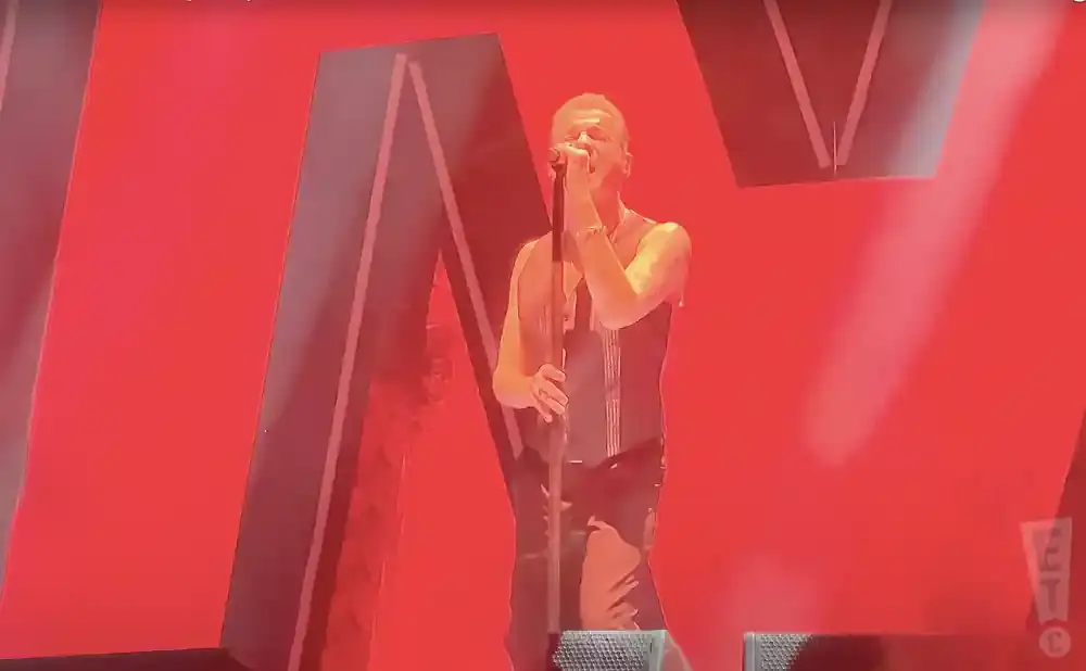 A man in a black vest sings into a microphone on a stage with a red background.