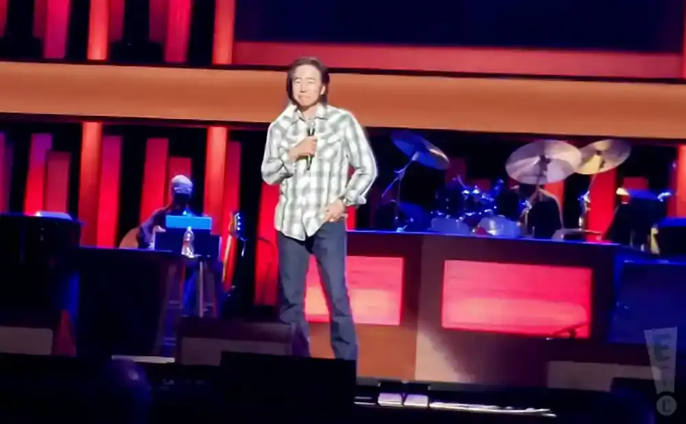 A man in a plaid shirt stands center stage with a microphone, while a drummer plays in the background.