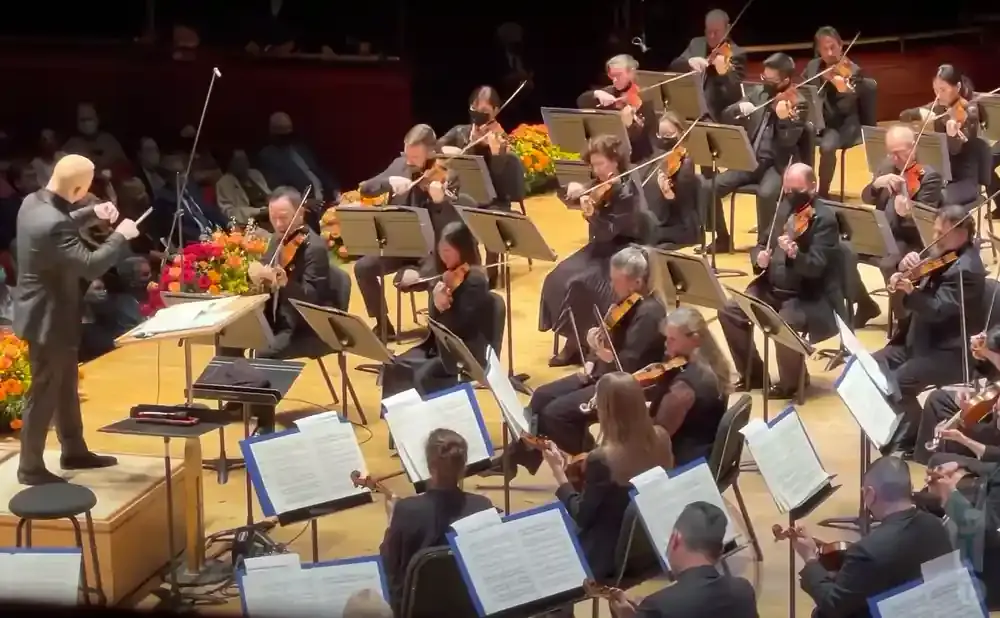 An orchestra conductor directs musicians playing violins and other instruments on stage.