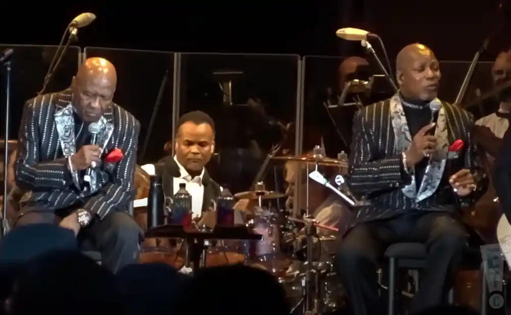 Three Black men in sparkly suits perform on stage. Two sing into microphones, one plays drums.