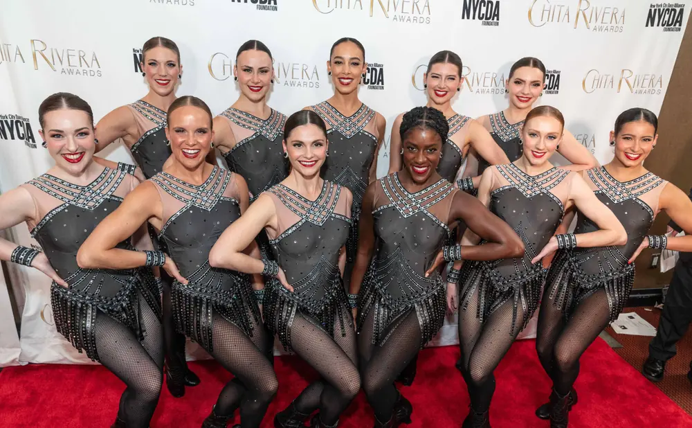 Dancers in sparkly black outfits pose on a red carpet in front of a 