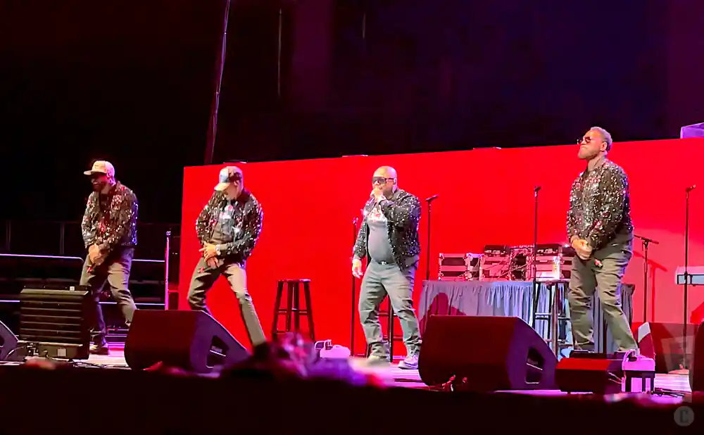 Four men on stage wearing sparkling jackets and hats, performing in front of a red backdrop.